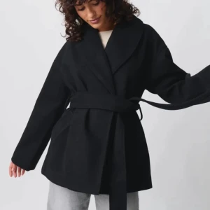 Svart kappa med bälte och bred krage - Stilren svart kappa med bred krage och knytskärp i midjan. Jackan har en rak och lite oversized passform med långa ärmar. Perfekt för dig som gillar en clean och klassisk look. Materialet ser ut att vara ull eller en ullmix.