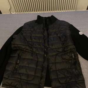 Svart Moncler pufferjacka med dragkedja - Säljer en svart pufferjacka från Moncler med quiltad front och ärmar i stickad kvalitet. Jackan har två fickor med dragkedja, hög krage och klassisk Moncler-logga på ärmen. Perfekt för dig som vill ha en stilren och exklusiv look.