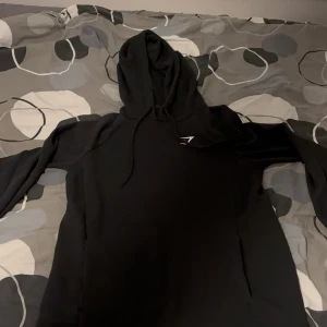 Svart hoodie från Gymshark S - Svart hoodie från Gymshark i storlek S. Tröjan har huva med snörning och en liten vit logga på bröstet. Tillverkad i en mjuk bomulls- och polyesterblandning med lite elastan för extra komfort. Perfekt för chill eller träning.