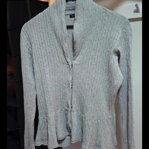 Grå ribbad kofta från Mywear, M - Supersöt grå kofta från Mywear med ribbat mönster, volangkant nedtill och knappar framtill. Långärmad och med en lite högre krage som ger en chill men ändå snygg look. Perfekt att slänga över en topp när det blir kyligt.