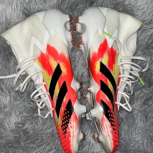 Adidas Predator 20.1 fotbollsskor - Säljer ett par Adidas Predator 20.1 fotbollsskor i storlek 44 2/3. Skorna har en cool design med vit bas och röda, gula och svarta detaljer. De har snörning, platt sula med dobbar och en strumpliknande form för extra stöd. Perfekta för dig som vill sticka ut på planen.
