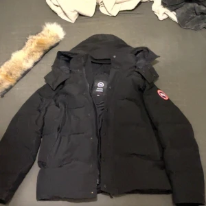 Svart dunjacka från Canada Goose S - Säljer en svart Canada Goose dunjacka i modell Fusion S/P. Jackan har huva med avtagbar pälskant, dragkedja och knappar framtill samt klassisk logga på ärmen. Perfekt för kalla vinterdagar och riktigt snygg streetstil.