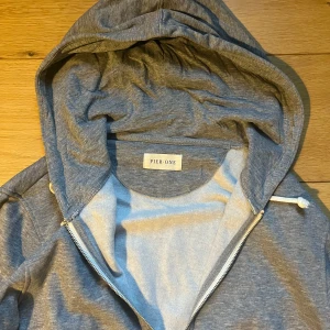 Grå hoodie med dragkedja från Pier One - Säljer en klassisk grå hoodie från Pier One med dragkedja framtill och vit dragsko i huvan. Perfekt för en avslappnad stil och enkel att matcha med det mesta. Mjuk och bekväm bomullsblandning.