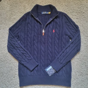 Marinblå stickad tröja Polo Ralph Lauren - Säljer en marinblå kabelstickad tröja från Polo Ralph Lauren med halv dragkedja och läderdetalj. Tröjan har lång ärm, ribbade muddar och röd broderad logga på bröstet. Perfekt för lager-på-lager under kyliga dagar. Accepterar bud.