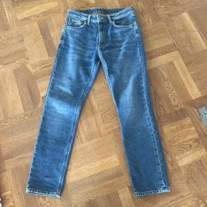 Blå jeans från Nudie Jeans Co, 27/28 - Säljer ett par klassiska blå jeans från Nudie Jeans Co i storlek 27/28. Jeansen har raka ben är slim, fem fickor och snygga kontrastsömmar. De är tillverkade i slitstarkt denim och har Nudie-loggan på bakfickan. Perfekta för en avslappnad och stilren look.