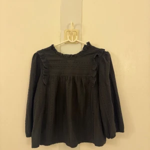 Svart blus med volang från JDY - Supersöt och svart blus från JDY med volangdetaljer vid axlar och halsringning. Blusen har lång ärm och är har design med små hålmönster. Perfekt för dig som gillar romantiska och feminina detaljer.