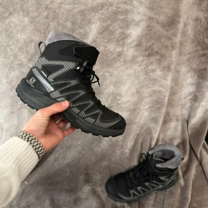 Svarta Salomon kängor med grå detaljer - Salomon kängor i svart med grå och vita detaljer, robust sula med grovt mönster för extra grepp. Skorna har snörning och är tillverkade i syntetmaterial med vadderad insida för extra komfort. Perfekta för utomhusaktiviteter under kyligare säsonger. Har använts bara 2 ggr.