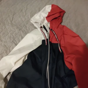Tommy Hilfiger vårjacka/ 2 Windbreaker Storlek XS - Snygg Tommy Hilfiger vindjacka/ vårjacka  storlek XS  i klassiska färger – röd, vit och marinblå. Den är Lätt väger jätte lite och vindtät och vattentät perfekt för vår och höst.   Har huva, dragkedja och dragsko i nederkant. Och två fickor med  Bekväm, stilren  fint skick! Den finns i Linköping men kan skickas med post! Men då måste mottagaren betala för frakten dock!