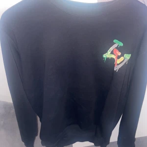 Lv tröja - Svart långärmad tröja från Louis Vuitton med stort färgglatt LV-tryck på ryggen och mindre tryck på bröstet. Tröjan har rund hals och är tillverkad i mjukt bomullsmaterial. Perfekt statement piece för dig som vill sticka ut.