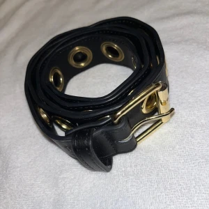Bälte - Snyggt svart bälte med stora guldfärgade öljetter längs hela bandet och matchande guldfärgat spänne. Perfekt accessoar för att ge outfiten en edgy vibe. Bältet är brett och har en cool, modern look. Inga defekter