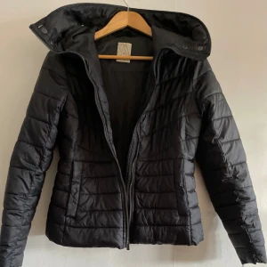 Svart lättviktsjacka (avtagbar pälsluva)Etirel S - Säljer en svart quiltad pufferjacka från Etirel med avtagbar huva och dragkedja framtill. Jackan har snygga horisontella sömmar, två fickor och en fuskpälsdetalj på huvan. Perfekt för kyliga dagar och riktigt skön passform.