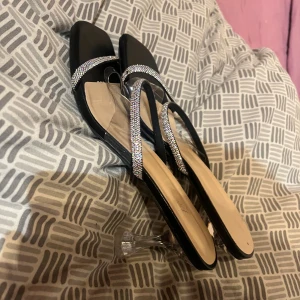 Svarta sandaler med glittriga remmar - Snygga svarta sandaler med fyrkantig tå och transparent klack. Remmarna är dekorerade med glittriga stenar som ger extra bling. Perfekt för dig som gillar att sticka ut med stilrena detaljer och en modern look.aldirg används 