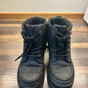 Svarta boots med snörning - Säljer ett par svarta boots med snörning och platt sula. Skorna har rund tå, grov yttersula och vadderad krage i textil för extra komfort. Materialet ser ut att vara syntet med mockakänsla och insidan är fodrad för värme. Perfekt för höst och vinter.