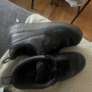 Svarta Nike Air Force 1 sneakers - Säljer ett par svarta Nike Air Force 1 sneakers med lågt skaft. Skorna har klassisk rund tå, svart skinn och textil på swoosh-loggan samt svart snörning. Sulan är grå och det finns en liten blå detalj på sidan. Perfekta för dig som gillar streetwear. Nypris 1700