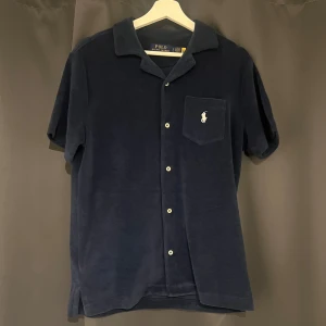 Mörkblå skjorta från Polo Ralph Lauren - Mörkblå skjorta i mjuk frotté från Polo Ralph Lauren med kort ärm, klassisk krage och vita knappar framtill. Bröstficka med broderad vit logga ger en clean och stilren look. Perfekt för dig som gillar avslappnad men snygg stil.