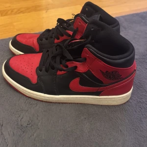 Nike Air Jordan 1 Bred Svart/Röd - Säljer ett par klassiska Nike Air Jordan 1 i färgen Bred med svart och rött skinn, vita mellansulor och röda yttersulor. Skorna har ikoniska Jordan Wings-loggan på sidan, snörning och perforerad tåbox. Perfekta för dig som vill sticka ut med en tidlös sneaker. 