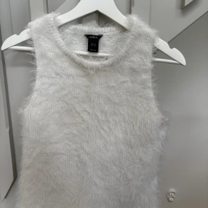 Vit fluffig topp från Lindex - Säljer en supermjuk och fluffig vit topp från Lindex. Toppen är stickad och har en ärmlös design med rund halsringning. Toppen är i storlek M men är liten i storleken, säljes för 100kr.