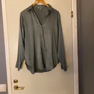 Grå silkeslen skjorta med knappar - Snygg grå skjorta från H&M i silkesliknande material. Skjortan har lång ärm, klassisk krage och knappar framtill samt vid manschetterna. Passformen är lite loose och tyget har en lätt glansig finish som ger en modern vibe.Frakt ingår inte 