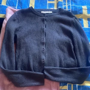 Mörkgrå ribbad kofta med knappar - Mysig mörkgrå kofta med ribbad struktur och knäppning framtill. Långärmad och i mjukt stickat material, perfekt att slänga på sig när det är kyligt. Enkel och stilren design som funkar till det mesta.