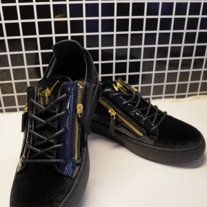 Giuseppe Zanotti - Säljer ett par svarta sneakers från Giuseppe Zanotti med lyxiga detaljer i guld, dragkedjor på sidorna och snörning framtill. Skorna har en mix av sammet och lack, samt platt sula och guldfärgad logga på plösen. Perfekta för dig som gillar statement-skor.