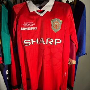 Manchester United Beckham 7 tröja - Legendarisk Manchester United fotbollströja från Champions League-finalen 1999 med Beckham och nummer 7 på ryggen. Röd med vita detaljer, broderat klubbmärke och SHARP sponsortryck. Klassisk krage och långärmad modell i polyester. OBS INTE ÄKTA
