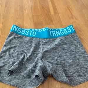 Grå sportshorts från H&M - Gråmelerade sportshorts från H&M med bred, turkos resår i midjan där det står TRNG8310. Shortsen är tighta och gjorda i ett stretchigt syntetmaterial, perfekta för träning och aktiva dagar.