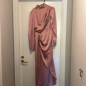 Rosa långklänning från SHEIN i M - Säljer en snygg rosa långklänning från SHEIN i storlek M. Klänningen har lång ärm, hög krage och cool drapering framtill. Materialet är glansigt och känns lätt mot huden. Dragkedja i ryggen och v-ringning framtill för extra stilpoäng.
