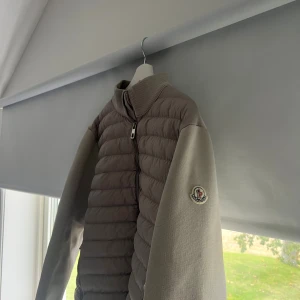 Beige  Moncler cardigan - Snygg beige pufferjacka från Moncler med quiltad framsida och stickade ärmar. Jackan har hög krage, två fickor med dragkedja och Moncler-logga på ärmen. Perfekt för dig som vill ha en clean och stilren look med exklusiv känsla. Passar L och xl, och med en hoodie under även M.