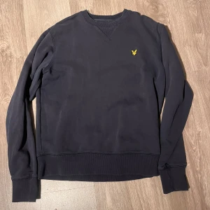 Svart sweatshirt från Lyle & Scott - En stilren svart sweatshirt från Lyle & Scott med deras klassiska gula logga broderad på bröstet. Tröjan har rund hals, ribbade muddar och är tillverkad i 100% bomull. Perfekt för dig som gillar enkel och snygg streetstil.