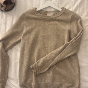 Beige stickad tröja från Vila - En klassisk beige stickad tröja från Vila med rund halsringning och ribbade muddar vid ärmslut och nederkant. Perfekt för lager-på-lager och enkel att matcha med jeans eller kjol. Mjuk och skön känsla, passar dig som gillar stilren och enkel design.