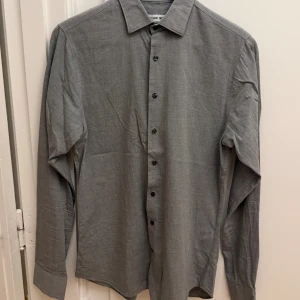 Grå skjorta från The Shirt Factory - Snygg grå skjorta från The Shirt Factory i slim fit-modell. Skjortan är tillverkad i 100% bomull, har klassisk krage, långa ärmar och knäppning framtill med svarta knappar. Perfekt för dig som gillar stilren och enkel design. Storlek 38.