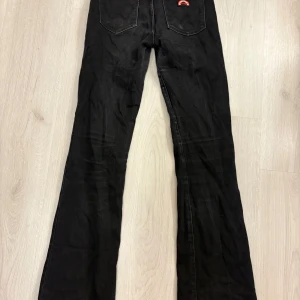 Svarta flare jeans från Wrangler - Svarta jeans från Wrangler i modellen Flare med utsvängda ben och klassiska fickor både fram och bak. Snyggt svart tyg med kontrastsömmar och Wrangler-logga på bakfickan. Perfekta för dig som gillar retrovibbar och vill ha en cool siluett.