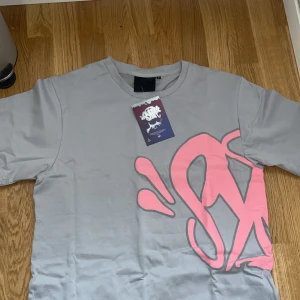 Syna wrld tshirt - Snygg grå t-shirt från SNAK med stort rosa graffiti-inspirerat tryck som täcker både fram- och baksidan. Klassisk rund hals och korta ärmar. Tillverkad i mjuk bomull för en skön känsla. Perfekt för dig som gillar streetwear och unika prints. S men passar M