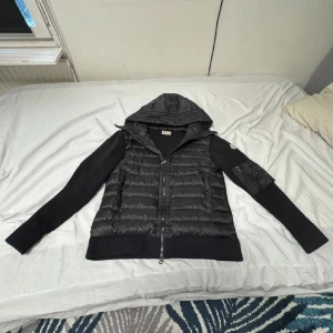 Svart Moncler cardigan med huva - Svart cardigan från Moncler med stickade ärmar och vadderad kropp. Jackan har huva, dragkedja framtill och en liten ficka med dragkedja på ena ärmen. Klassisk Moncler-logga på ärmen och diskret glansig finish på det vadderade partiet. Använd runt 2 gånger men säljer för den va för lite.