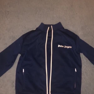 Blå track jacket från Palm Angels - Snygg marinblå track jacket från Palm Angels med vita ränder längs ärmarna och dragkedja framtill. Jackan har hög krage, två fickor med dragkedja och logga på bröstet. Perfekt för en sportig och trendig look.