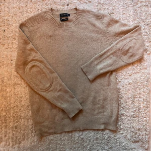 Beige stickad tröja från East West - Beige stickad tröja från East West med rund halsringning och ribbade muddar. Tröjan har förstärkta armbågslappar och en clean, klassisk look som passar perfekt till jeans eller chinos. Skön och mjuk känsla tack vare det stickade materialet. Men har en liten fläck på höger arm men syns inte jättemycket. 