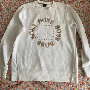 Vit sweatshirt från Hugo Boss - Vit sweatshirt från Hugo Boss med rund hals och tryck i beige och brunt framtill där det står 'BOSS' i en cirkel. Tröjan har långa ärmar, ribbade muddar och är gjord i mjuk bomull. Perfekt för en clean och stilren look.  Storlek S men passar också M väldigt bra , skulle säga en stor S tröja. Ordinarie pris 1400 kr.