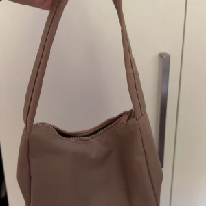 Beige axelväska med dragkedja - Väskan har en enkel, modern design och är tillverkad i ett mjukt syntetmaterial. 