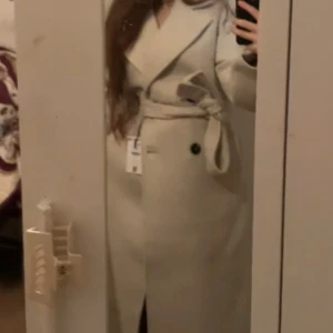 Beige kappa från Bershka med bälte - Stilren beige kappa från Bershka med bred krage, knytskärp i midjan och svarta knappar. Jackan har långa ärmar och klassisk passform, perfekt för dig som vill ha en clean och trendig look. Materialet är mjukt och känns lyxigt. ((Köpt för 800 något minns ej, användbar men är i bra skick för och vara använd i ett år.))