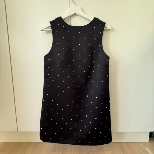 Svart kort klänning med paljetter på och en rosett i ryggen - Supersöt svart ärmlös klänning med paljetter på och en snygg v-ringning bak som pryds av en liten rosett. Klänningen är kort och har en rak, enkel passform. 