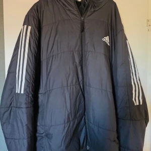 Svart pufferjacka från Adidas XL - Svart pufferjacka från Adidas i storlek XL. Jackan har klassiska vita ränder längs ärmarna och Adidas-logga på bröstet. Dragkedja framtill och oversized passform. Perfekt för kalla dagar och sportig stil.