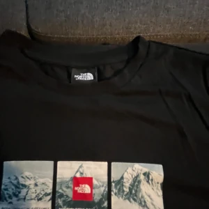 Svart t-shirt The North Face med tryck - Svart t-shirt från The North Face med coolt tryck av berg i tre rutor på bröstet, där mittenrutan har röd logga. Klassisk passform och rund hals. Perfekt för dig som gillar streetwear och outdoor vibes.