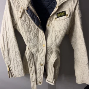  beige jacka från Barbour - beige jacka från Barbour International med bälte i midjan, guldiga knappar och dragkedja. Jackan har lång ärm, bröstficka med Barbour-logga. Jackan har inga tecken på användning.