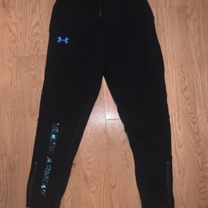 Under armour träningsbyxor - Svarta träningsbyxor från Under Armour med blå logga på låret och glansig text längs benet. Byxorna har elastisk midja, dragsko och dragkedjor vid bensluten. Lätta och sköna med loose fit, perfekta för träning eller chill.