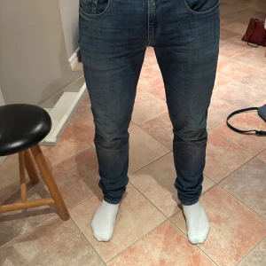 Blå Replay Anbass jeans slim fit 34/34 - Säljer ett par blå Replay Anbass jeans i storlek 34. Slim fit-modell med klassiska fem fickor, snygga slitningar och detaljerad söm på bakfickorna. Jeansen har normal midja och är tillverkade i mjukt denimtyg för en skön passform. Använt ett fåtal gånger