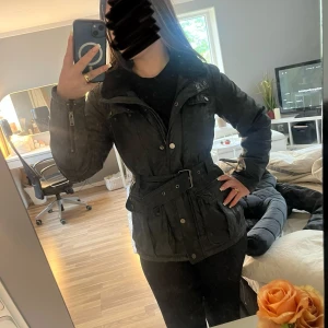 Svart parkajacka från Stockh LM med bälte - Snygg svart parkajacka från Stockh LM med fuskpäls på kragen, bälte i midjan och flera fickor framtill. Jackan har dragkedja, knappar och detaljer i silver. Perfekt för kyliga dagar och har en cool, urban vibe. Original pris är runt 1500-2000 😊