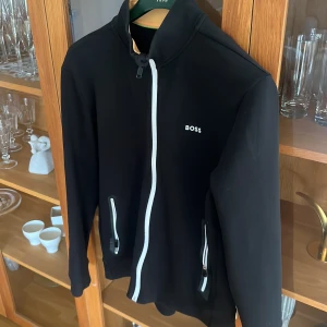 Svart ziptröja från BOSS - Svart långärmad ziptröja från BOSS med vit dragkedja och logga på bröstet. Tröjan har två fickor med dragkedja och en stilren, sportig look. Perfekt för dig som gillar enkel och clean design.