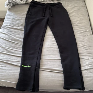 Svarta mjukisbyxor syna med grön print - Svarta mjukisbyxor från syna med grön graffiti-print på baksidan och broderad text 'Forgive Me' på benet. Byxorna har snörning i midjan, dragkedjor vid bensluten och är gjorda i mjukt bomullsmaterial. Perfekta för chill eller streetwear.