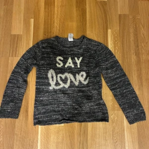Grå stickad tröja med text från C&A - Mysig gråmelerad stickad tröja från C&A med vit text 'SAY love' framtill. Tröjan har lång ärm, rund hals och är tillverkad i en blandning av akryl, bomull, polyester och metalliserat garn för en cool glansig effekt.
