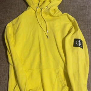 Gul hoodie från Calvin Klein Jeans XS - Säljer en riktigt snygg gul hoodie från Calvin Klein Jeans i storlek XS. Tröjan har huva med snörning, stor CK-logga på ärmen och en klassisk känguruficka framtill. Perfekt för dig som vill sticka ut med färg och streetstyle.